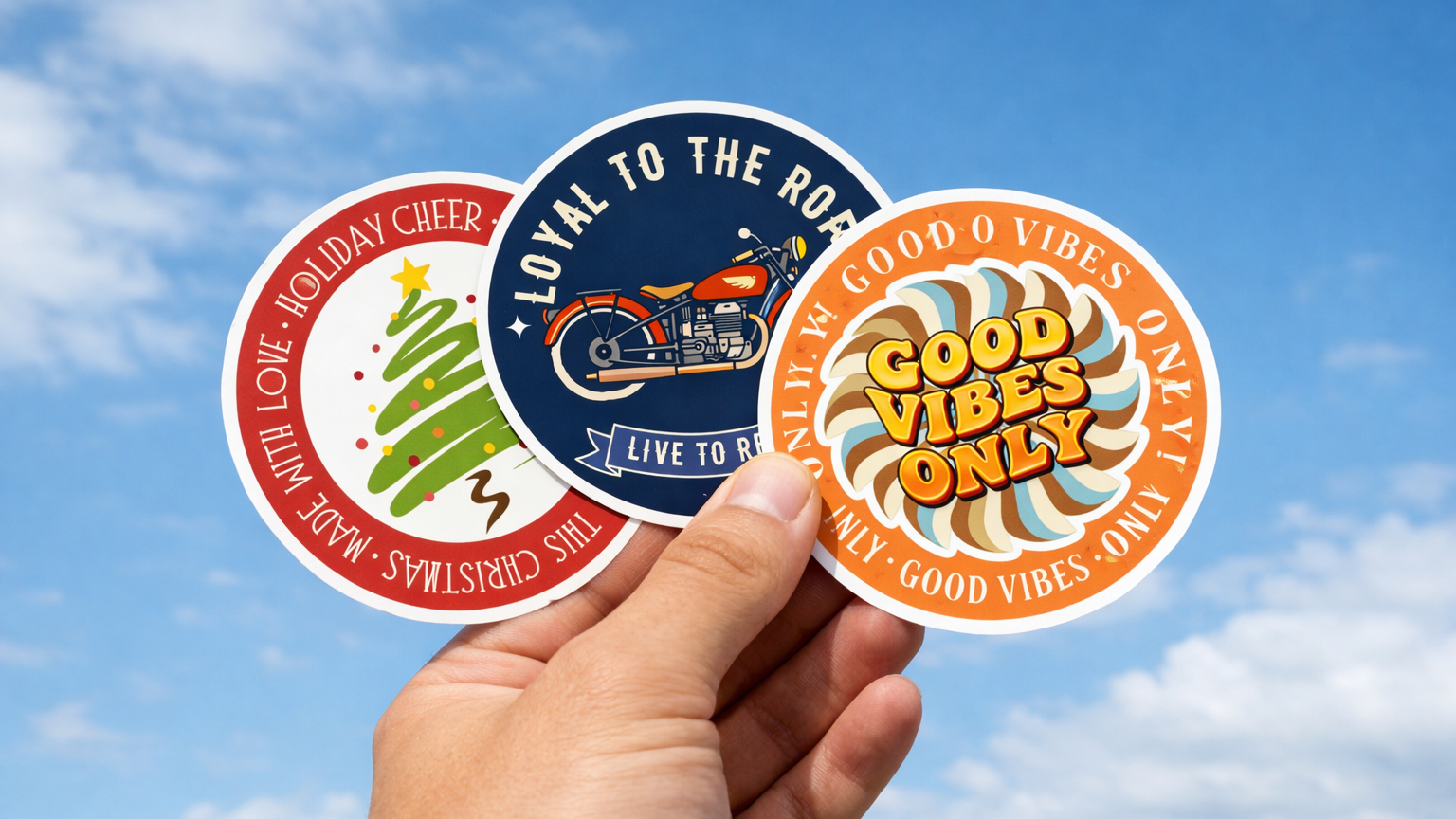 Hand holding three perfect custom vinyl sticker examples against a blue sky – Christmas tree design, "Loyal to the Road" motorcycle design, and "Good Vibes Only" design – for a guide on how to order custom stickers