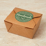 A 2-inch round green "Handmade" label applied to seal a brown kraft paper food box. This 2-inch circle sticker demonstrates professional packaging for artisan quality goods.