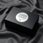 A round silver foil brand label with "Curated Necklaces" text and jewelry icon, applied to a luxury black gift box against a dark silk fabric background.