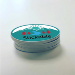 A stack of custom oval stickers with a teal and white mountain design, featuring red pine trees, a winding river, and a rising sun. The brand name "Stickable" is printed in bold black text at the bottom.