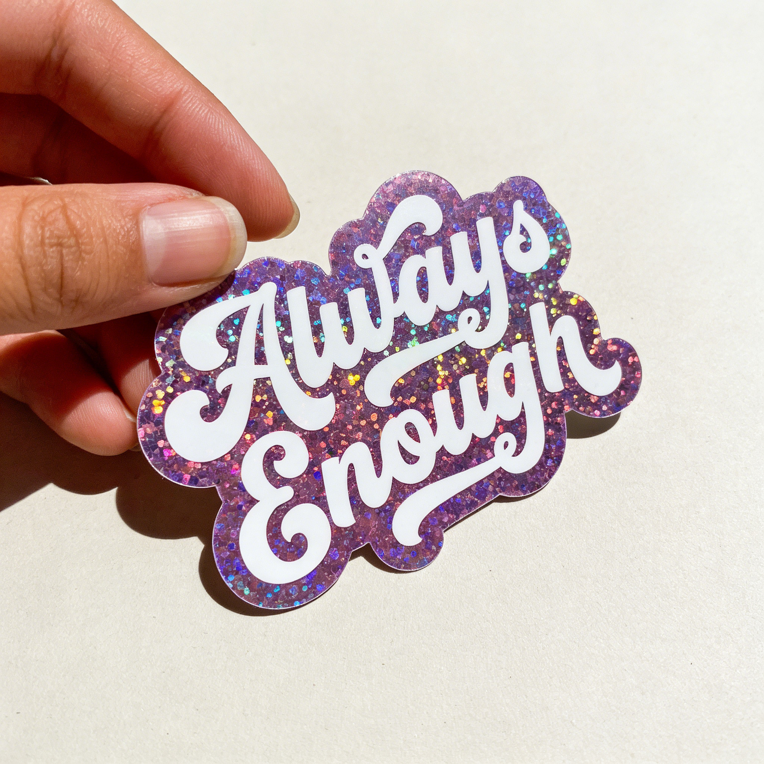 Hand holding a glittery 'Always Enough' sticker against a plain background