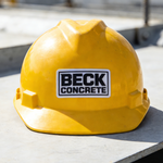 Yellow hard hat with Beck Concrete custom sticker on a construction site