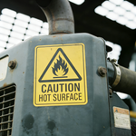 Close-up of a machine with a “Caution Hot Surface” warning sticker made from industrial-grade vinyl, background softly blurred.