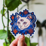 Sticker of a cat with celestial designs held by a hand against a natural background