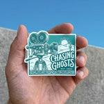 Hand-held view of a retro-style vinyl sticker with a teal background. The design includes a white line-art vintage movie camera on a tripod set against a desert landscape with a sun and cactus. Text reads 'Chasing Ghosts' in a stylized font with 'EST. 2024' below it.