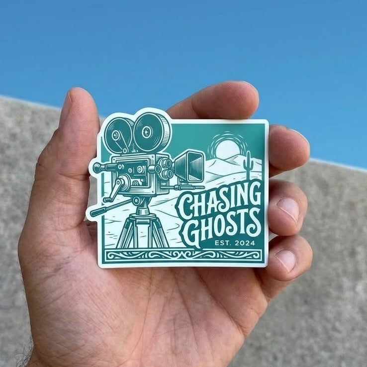 Hand-held view of a retro-style vinyl sticker with a teal background. The design includes a white line-art vintage movie camera on a tripod set against a desert landscape with a sun and cactus. Text reads 'Chasing Ghosts' in a stylized font with 'EST. 2024' below it.