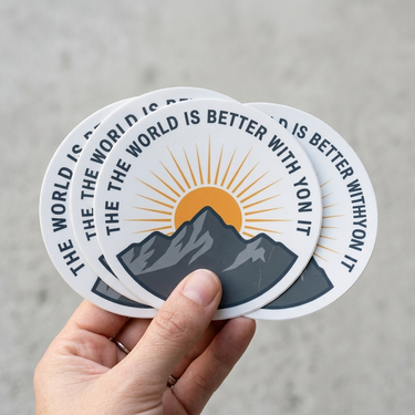Hand holding a stack of circular stickers with mountain and sun design on a gray background