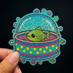 Colorful glitter sticker of a fish in a bowl held by a hand against a black background