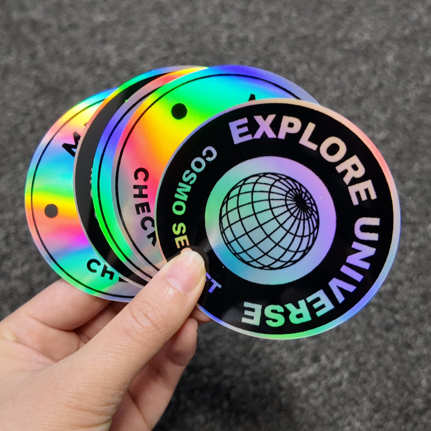 Hand holding a colorful circular holohgraphic stickers with 'Explore Universe' text on a dark background