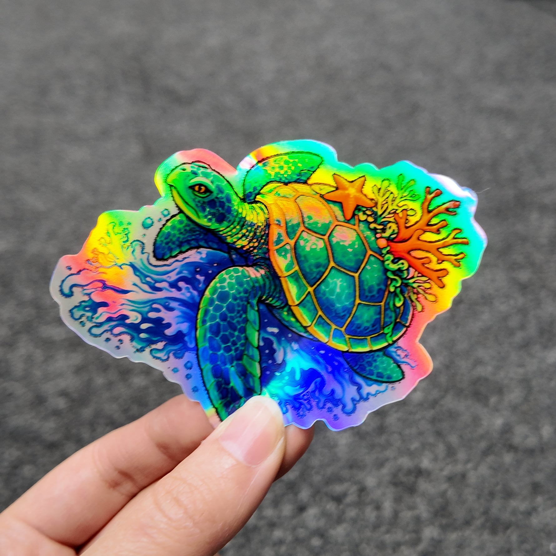 Colorful turtle and coral holographic sticker held by a hand against a dark background
