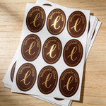 Custom oval sheet labels for handmade products, featuring brown and gold monogram stickers arranged on label sheets for boutique branding