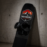Custom snowboard stickers featuring a mountain design on a black snowboard leaning against a concrete wall.