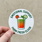Logo sticker with a drink illustration and text 'Emotional Support Farm Fresh Elixir' held by a hand on a beige background.