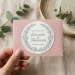 Hand holding a pink card with a circular wedding stickers featuring 'Eleanor & Theodore' on a textured surface with greenery.