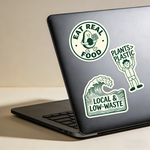 Laptop with eco themed stickers including “Eat Real Food”, “Plants Not Plastic”, and “Local & Low Waste” designs