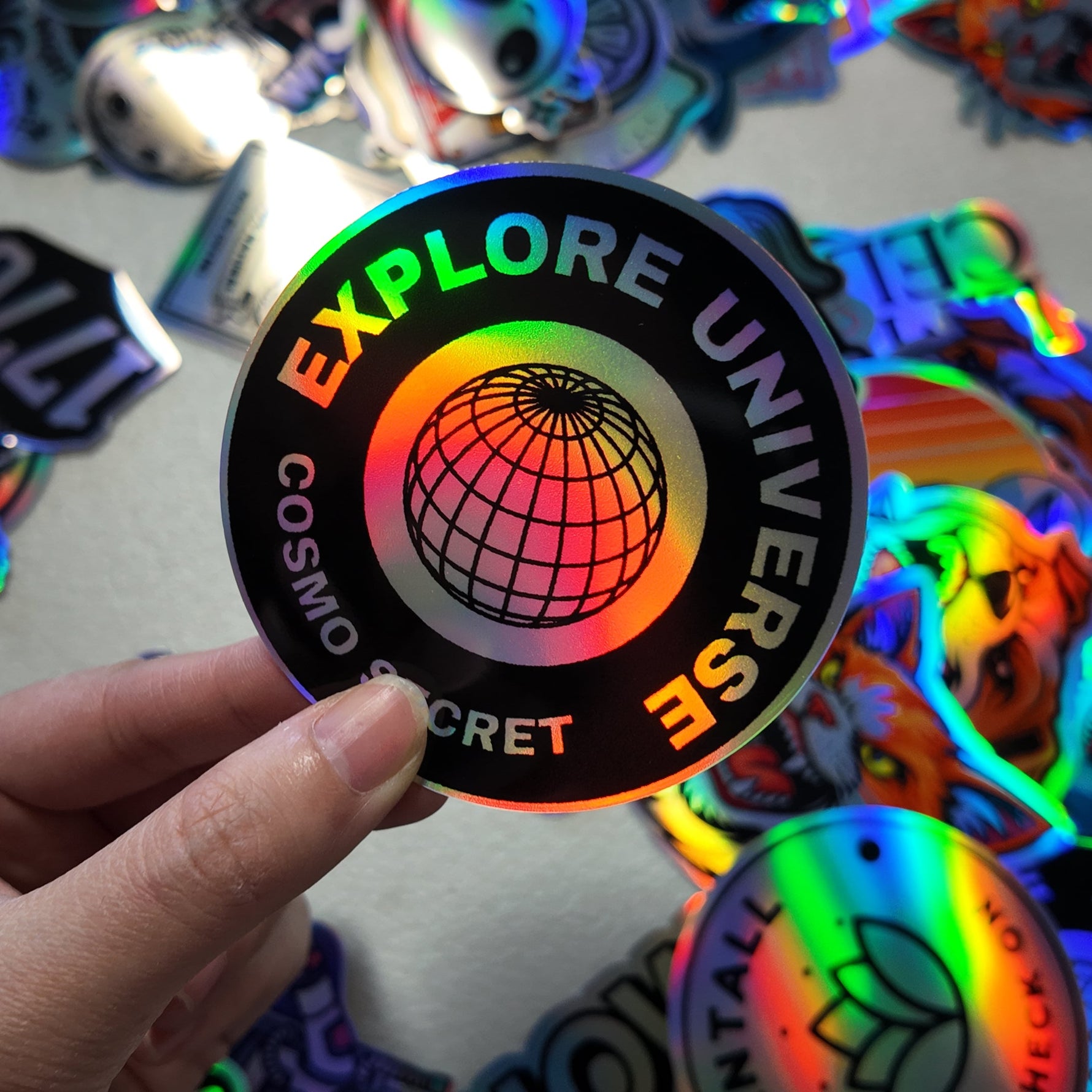 Hand holding a colorful 'Explore Universe' holographic sticker with more stickers in the background.