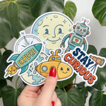 Four colorful stickers featuring a robot, submarine, rocket, and moon with 'Explore' and 'Stay Curious' text, held against a leafy green background.