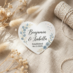 Heart-shaped decorative wedding stickers with names and date on a textured surface with dried flowers and twine.