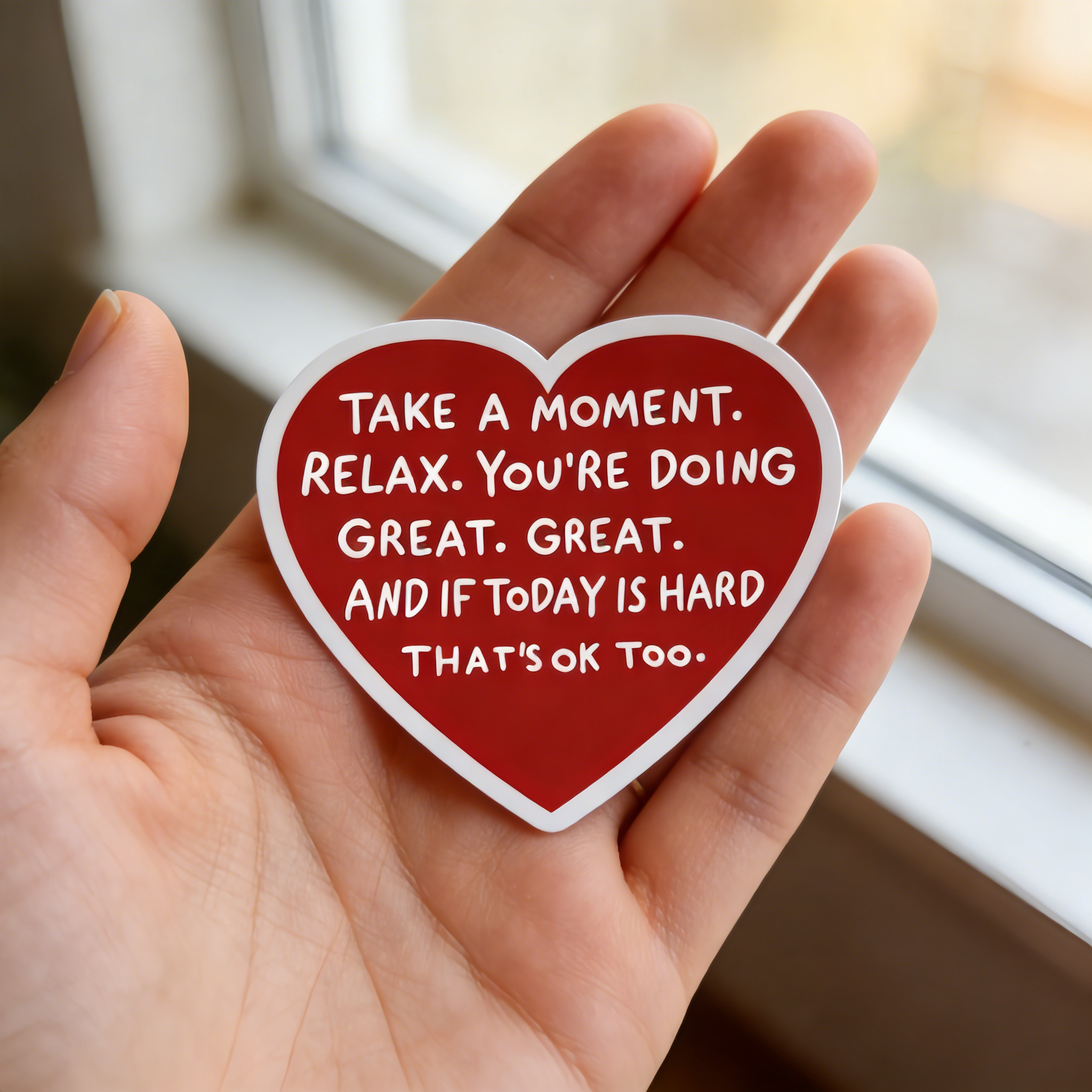 Heart-shaped sticker with motivational quote held in a hand by a window