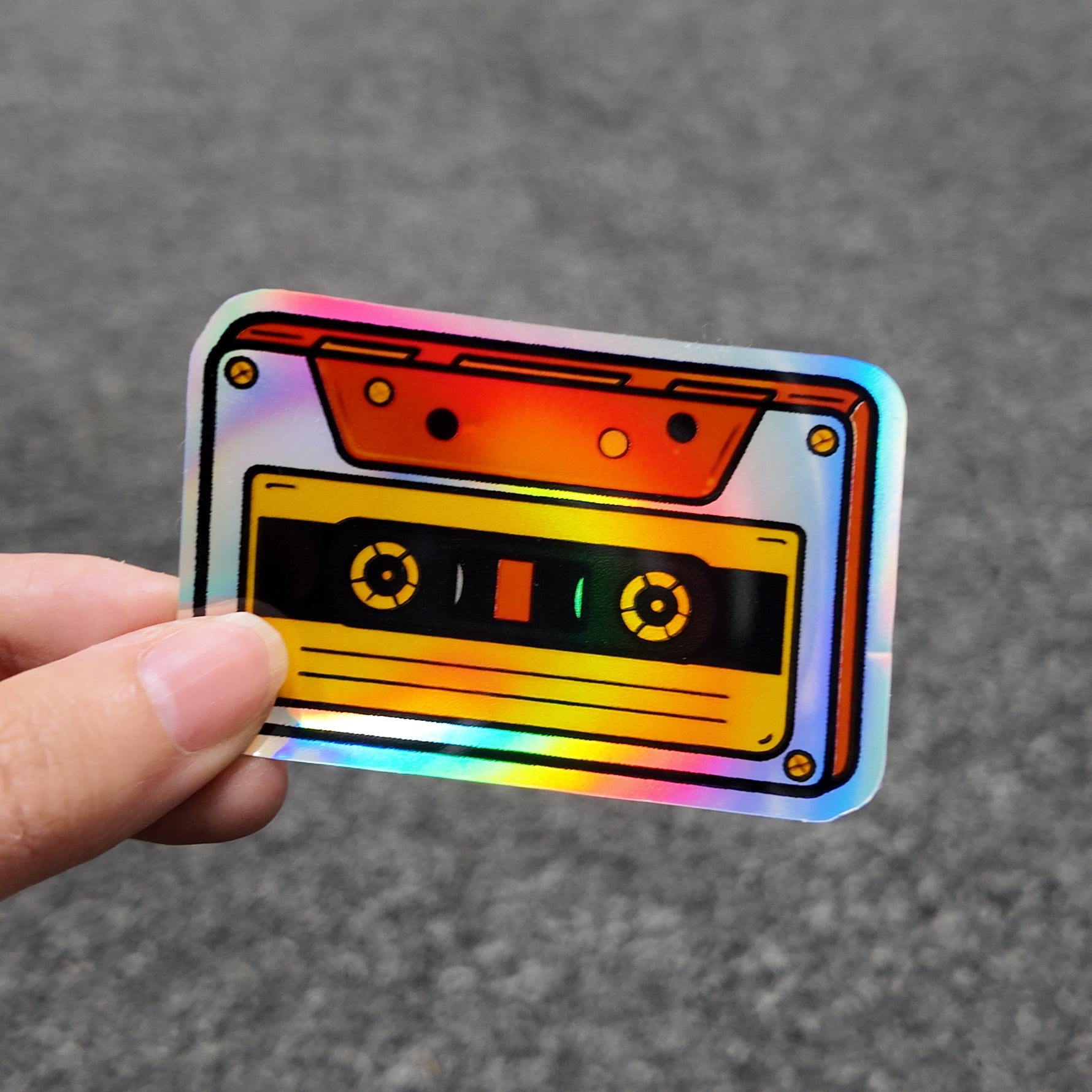 Holographic sticker of a cassette tape held by a hand on a gray background