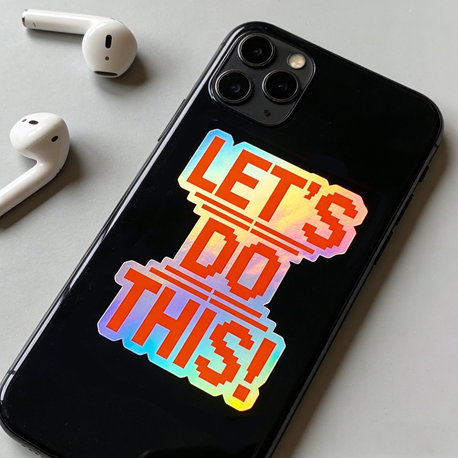 Holographic die cut vinyl sticker with pixel-style ‘LET’S DO THIS!’ text in rainbow reflective foil, applied on a black smartphone case on a gray desk with white wireless earbuds, showing an everyday use example