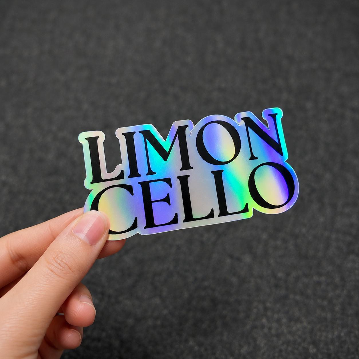 A hand holding a custom holographic sticker featuring the word "LIMONCELLO" in bold serif typography, printed on premium vinyl with a high-durability iridescent rainbow finish.