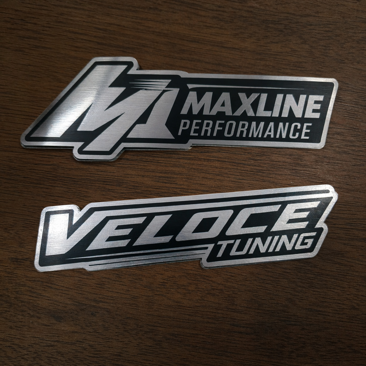Two automotive performance metallic silver vinyl stickers, one for Maxline Performance and one for Veloce Tuning, on a wooden surface.