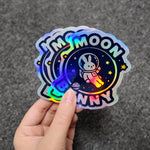 Moon and Bunny holographic sticker held by a hand on a gray background