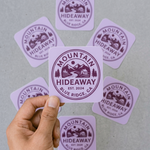 Hand holding a 'Mountain Hideaway' ounded corner sticker with multiple stickers for branding