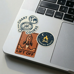 Laptop with custom stickers featuring a compass illustration with “Chart Your Course”, a pixel rocket “Lift Off” design, and a campfire “Campfire Nights” sticker
