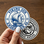 Two round stickers with Viking-themed designs held by a hand on a wooden surface.