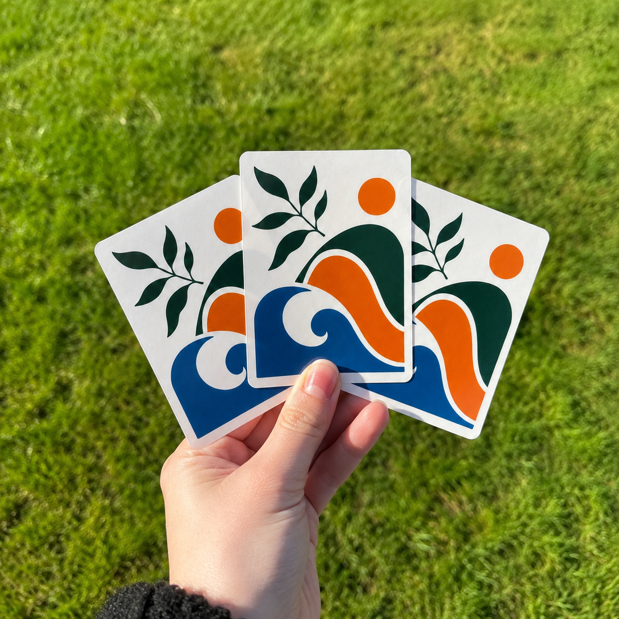 Hand holding three custom artistic vinyl stickers with abstract designs on a grassy background