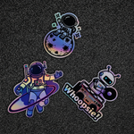Three colorful holographic stickers featuring an astronaut, a robot, and text on a dark textured background.