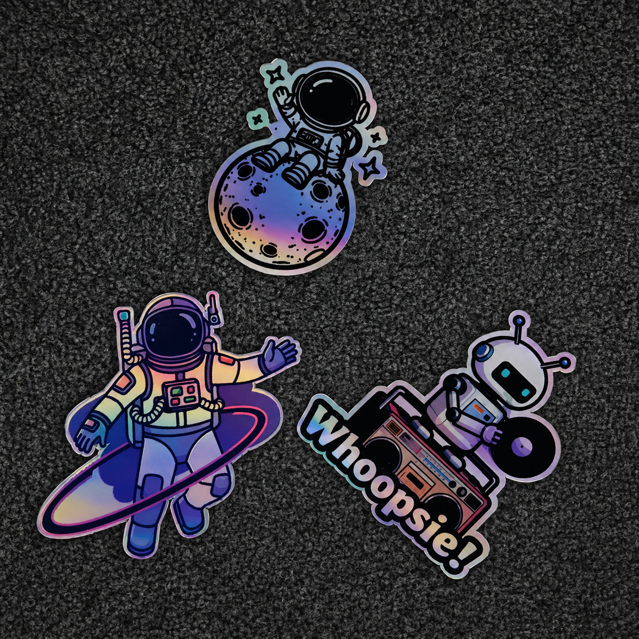 Three colorful holographic stickers featuring an astronaut, a robot, and text on a dark textured background.