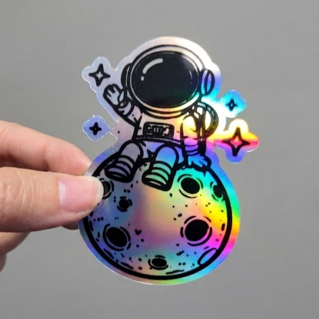 Hand-held view of a die-cut holographic vinyl sticker. The design features a cute astronaut character sitting on the moon and waving, surrounded by tiny sparkling stars. The material is dynamic with rainbow-colored light refraction.