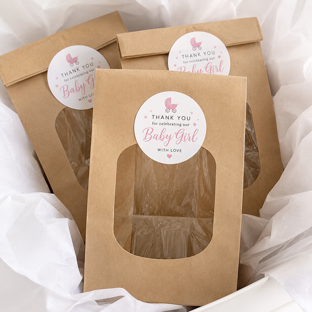 Three kraft paper gift bags with circle baby shower stickers 'Thank you for celebrating our Baby Girl with love,' arranged in white tissue paper.