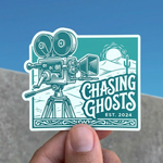 Sticker with a vintage camera design and 'Chasing Ghosts' text held by a hand against a blue sky.