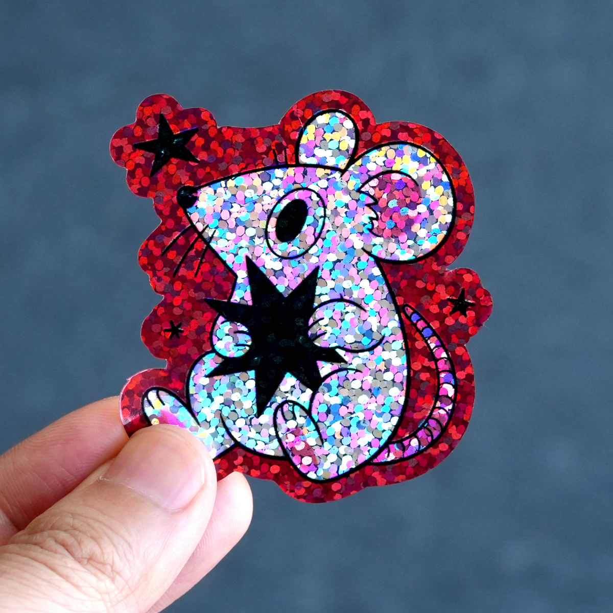 Hand holding a cartoon mouse glitter sticker with red sparkling accents on a dark blue background.