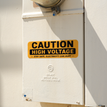 Yellow custom warning bumper sticker with CAUTION HIGH VOLTAGE text on an electrical box.