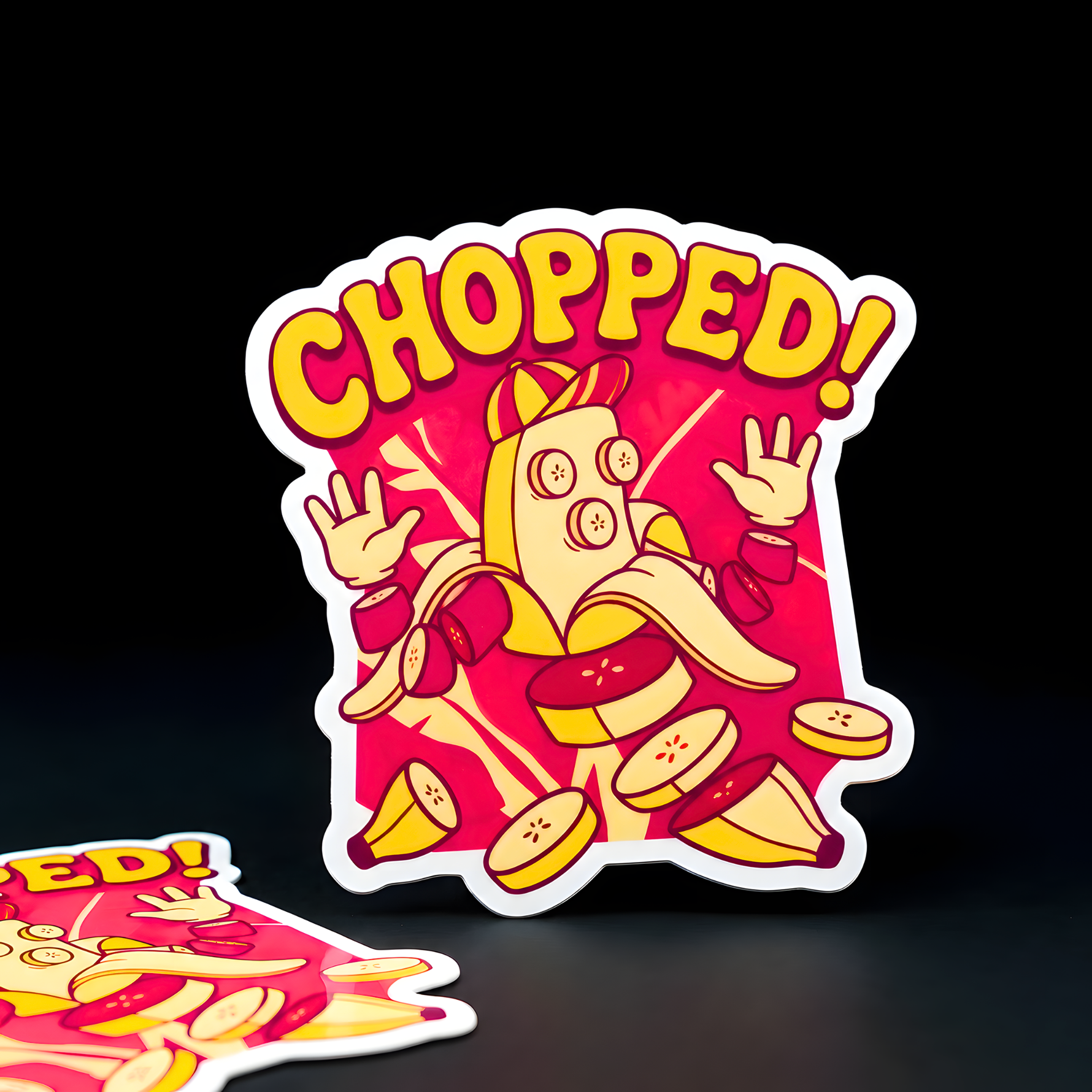 Die-cut vinyl sticker of a cartoon banana man being chopped into slices, vibrant yellow and deep pink design, professional full-volor printing.