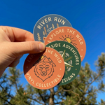 Four vinyl stickers with nature-themed designs held against a blue sky and tree background.