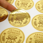 Close-up photograph of circular gold foil labels with an espresso machine design. A person's finger is gently lifting one custom gold label from the sheet, showcasing the metallic gold decal and reflective finish.