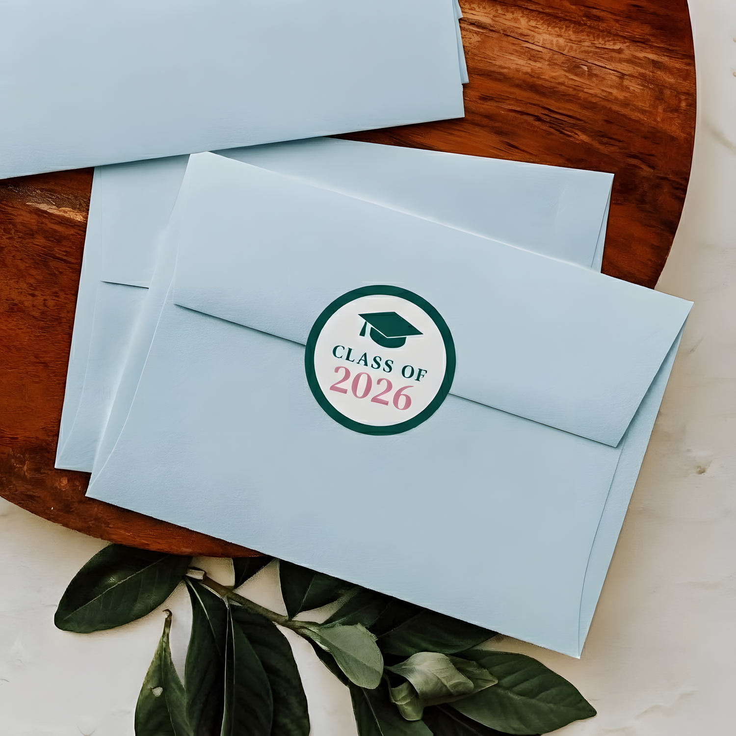 Light blue graduation invitation envelopes sealed with a custom Class of 2026 round sticker on a wooden tray.