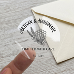 A round transparent personalized label with "Artisan & Handmade" text and a lavender illustration used as a decorative seal on a cream envelope.