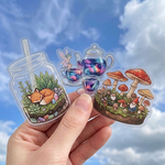Hand holding three colorful illustrated clear stickers made of transparent vinyl with full-color printing against a blue sky with clouds.