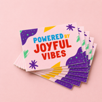 Colorful rectangle stickers with Powered by Joyful Vibes slogan stacked on a pink background