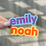 Colorful Emily and Noah name stickers held by hand near a window.
