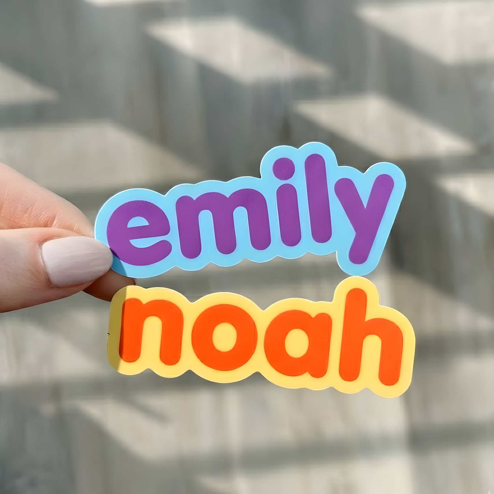 Colorful Emily and Noah name stickers held by hand near a window.