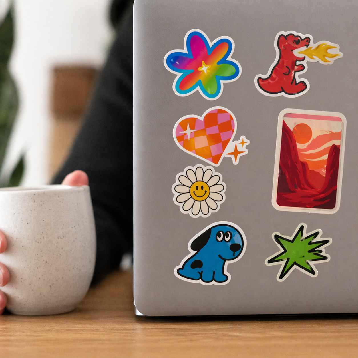 macbook pro laptop with colorful stickers on a wooden table, person holding a mug.