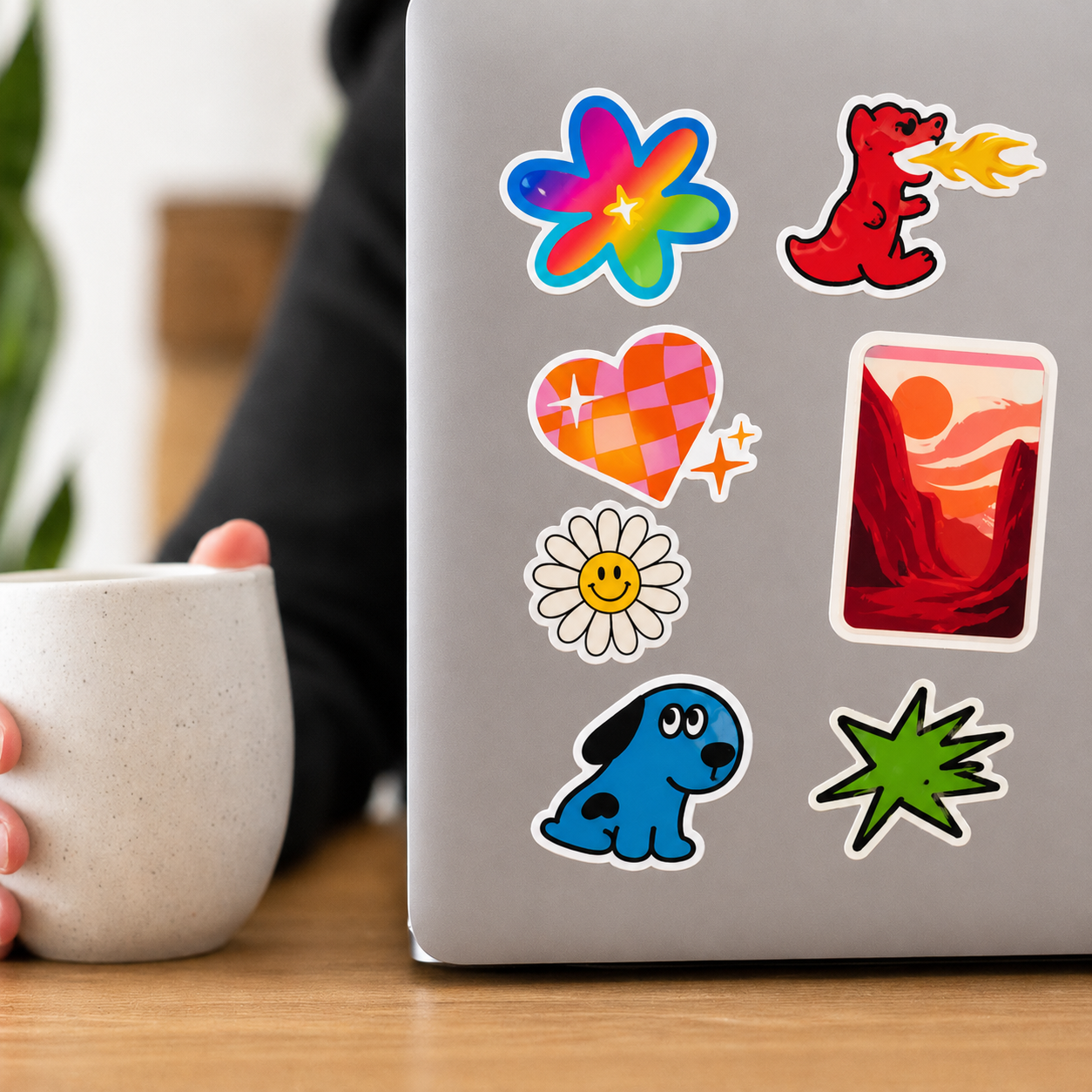 macbook pro laptop with colorful stickers on a wooden table, person holding a mug.